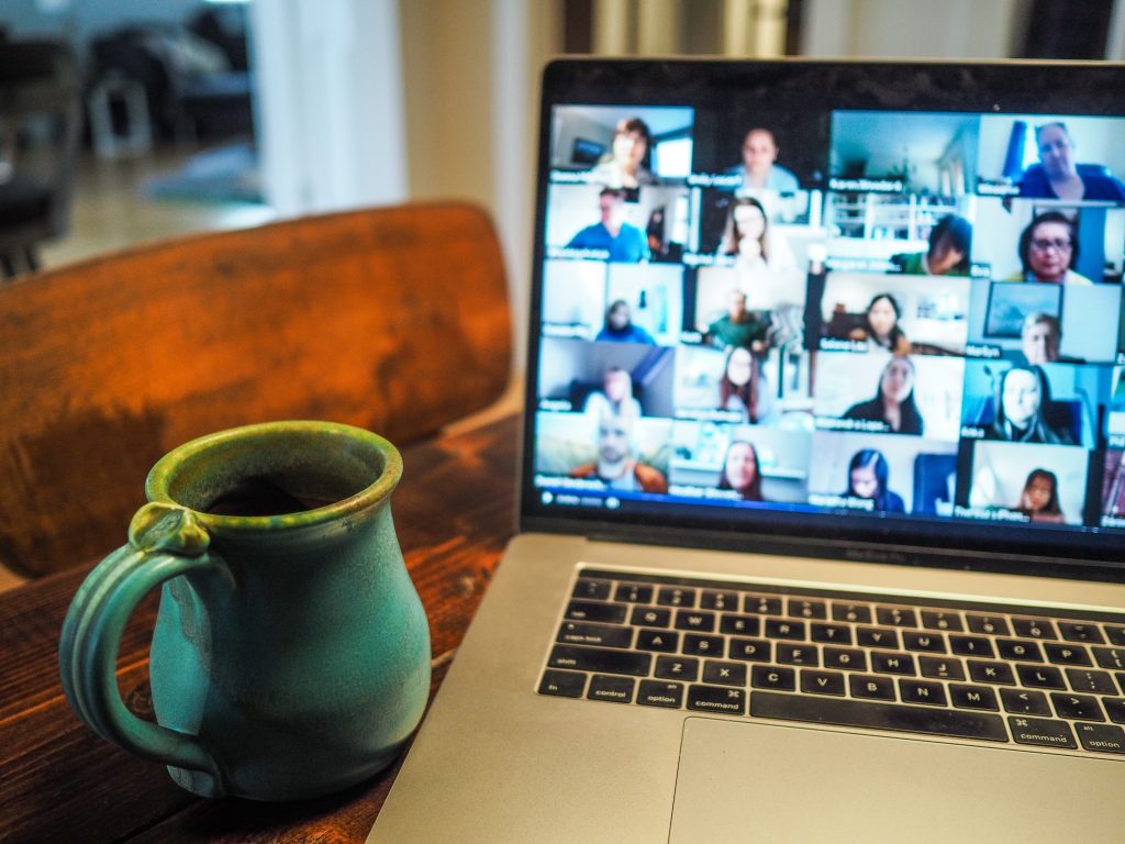 A laptop opened on a Zoom screen showing various participants on screen. A green mug sits next to the laptop, which is sitting on a wooden table, next to a wooden chair